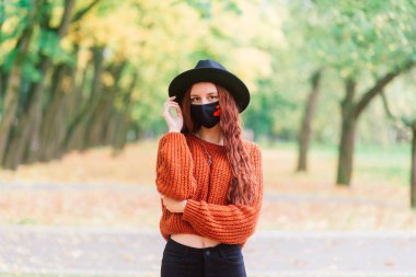 Sarı sonbahar parkında siyah stil maske takan kırmızı kazaklı genç bir kadının portresi. Moda, yaşam tarzı, karantina, koronavirüs..