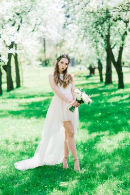 Beyaz gelinlikli gelin yeşil parkta bir buket tutuyor. Güneşli bir günde yaz düğünü..