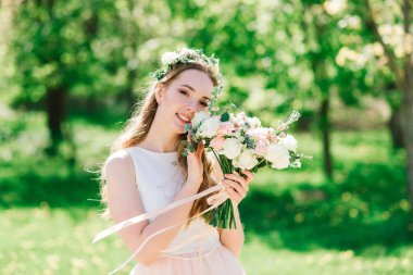 Beyaz gelinlikli gelin yeşil parkta bir buket tutuyor. Güneşli bir günde yaz düğünü..