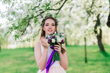 Beyaz gelinlikli gelin yeşil parkta bir buket tutuyor. Güneşli bir günde yaz düğünü..