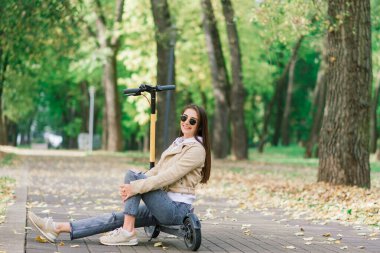 Sonbahar parkında elektrikli scooter kullanan genç bir kadın. Yeşil ulaşım, trafik sıkışıklığı.