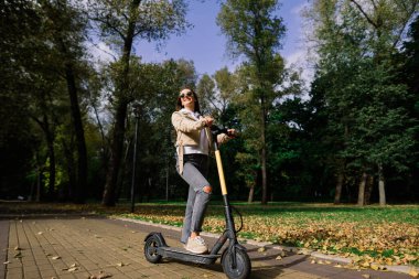 Sonbahar parkında elektrikli scooter kullanan genç bir kadın. Yeşil ulaşım, trafik sıkışıklığı.