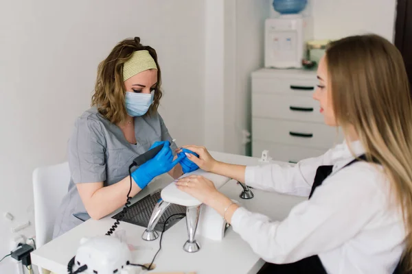 Manicure and pedicure salon, covid-19 and social distance. Master in rubber gloves and young woman client in protective mask in beauty studio interior.