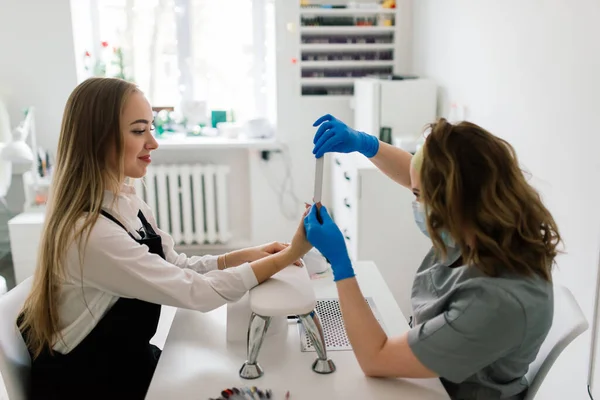 Manicure and pedicure salon, covid-19 and social distance. Master in rubber gloves and young woman client in protective mask in beauty studio interior.