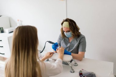 Manikürcü koruyucu maske takıyor. Manikür salonunda kadın tırnaklarını ojeyle boyuyor..
