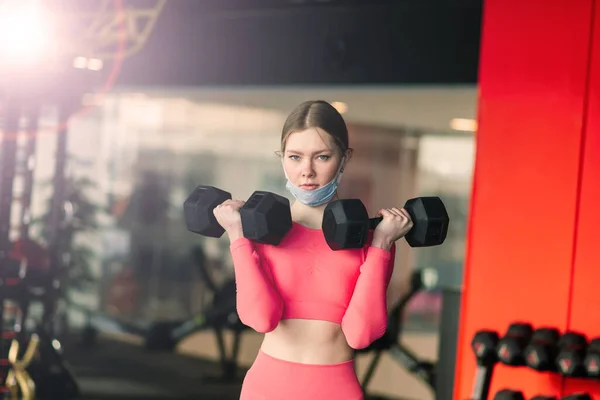 Female athlete fitness coach wearing white medical protective mask do exercises with dumbbell, invite to do fitness class in empty gym. Athletes against COVID-19