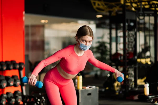 Female athlete fitness coach wearing white medical protective mask do exercises with dumbbell, invite to do fitness class in empty gym. Athletes against COVID-19