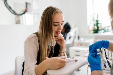 Güzellik salonundaki gülümseyen kadın manikür ustasından manikür yaptırıyor. Usta tırnak törpüsü kullanıyor.