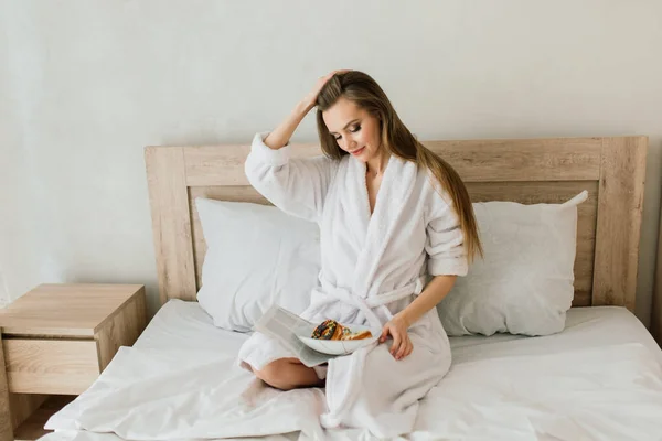 Young woman in white towel and robe in kitchen during quarantine. Hold donut on plate. Breakfast time at home.