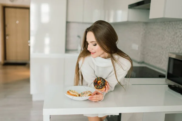 Young woman in white towel and robe in kitchen during quarantine. Hold donut on plate. Breakfast time at home.