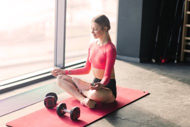 Bir kadın büyük bir pencereye karşı minder üzerinde yoga yapıyor.