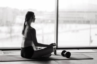 Bir kadın büyük bir pencereye karşı minder üzerinde yoga yapıyor.