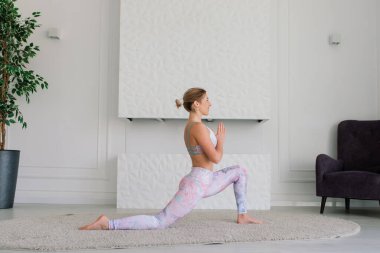 Genç, çekici, gülümseyen bir kadın yoga yapıyor, spor kıyafetleri giyiyor, sutyen giyiyor, evde eğitim görüyor.
