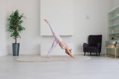 Genç, çekici, gülümseyen bir kadın yoga yapıyor, spor kıyafetleri giyiyor, sutyen giyiyor, evde eğitim görüyor.