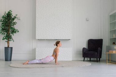 Genç, çekici, gülümseyen bir kadın yoga yapıyor, spor kıyafetleri giyiyor, sutyen giyiyor, evde eğitim görüyor.