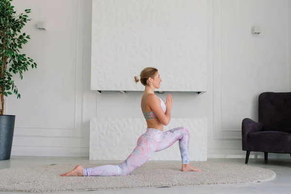 Genç, çekici, gülümseyen bir kadın yoga yapıyor, spor kıyafetleri giyiyor, sutyen giyiyor, evde eğitim görüyor.