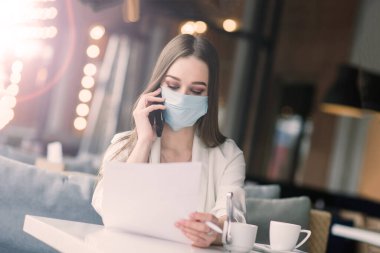 Koruyucu maskeli ciddi bir kadın bir kafede haberlere bakıyor.