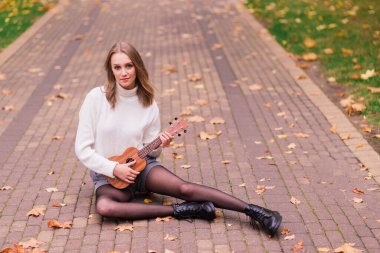 Genç bir kadın parkta bankta oturmuş gitar çalıyor ve gitar çalıyor.