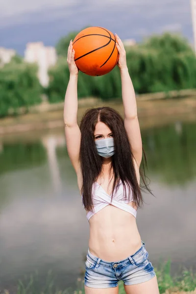 Young sport fitness woman basketball player using mask to protect with ball, lake background.