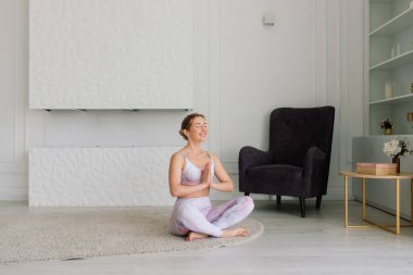 Genç, çekici, gülümseyen bir kadın yoga yapıyor, spor kıyafetleri giyiyor, sutyen giyiyor, evde eğitim görüyor.