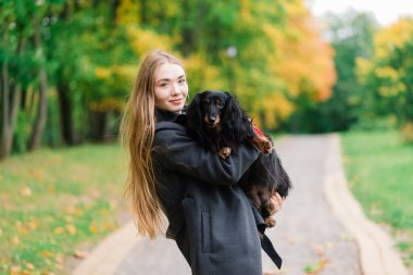 Sonbahar parkında küçük köpeğini kucağında tutan mutlu kadın.