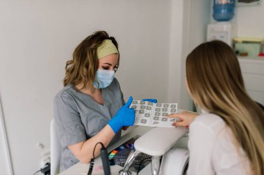 Manikürcü koruyucu maske takıyor. Manikür salonunda kadın tırnaklarını ojeyle boyuyor..