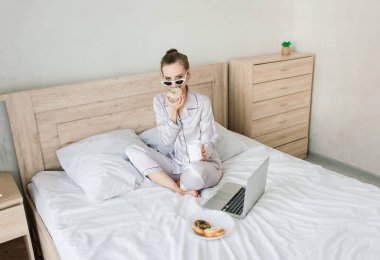 Pijama giymiş, dizüstü bilgisayarda yazan ve ışık odasında yatarken gülümseyen neşeli genç bir kadının fotoğrafı.