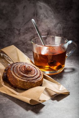 Cam bardakta çay kaşıkla, kağıt poşette çörek ile, gri noktalı arka planda...