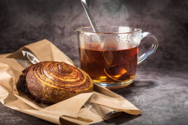 Cam bardakta çay kaşıkla, kağıt poşette çörek ile, gri noktalı arka planda...