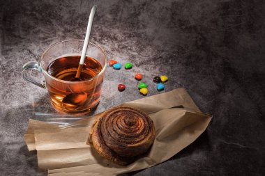 Cam bardakta çay, kaşık, kağıt torbada çörek ve gri renkli arka planda renkli şekerler.