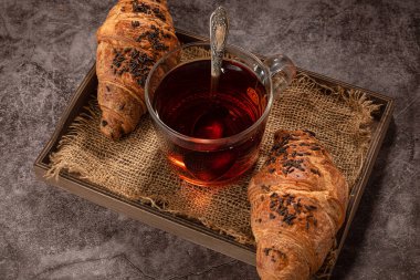 Çikolatalı kruvasan keki, şeffaf bir fincan çay ve kaşıkla, tahta tepside, koyu bir arka planda.