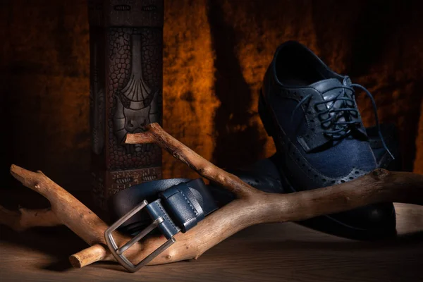 A man\'s belt of blue color, on a snag lying on an oak table top, against a background of burlap illuminated by an orange filter, a rectangular vase, and an inspector\'s shoe