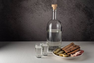 A glass bottle with glasses, with a strong drink and a snack on a white plate, pickles, bacon, bread, on a white countertop against a gray wall