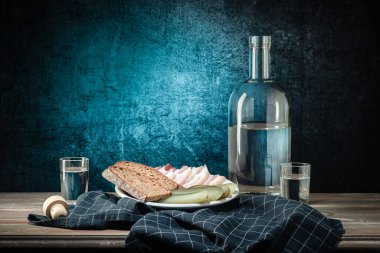 Bir şişe ve iki bardak, sert bir içki, ve beyaz bir tabak atıştırmalık, koyu bir peçete, arka planda bir leke