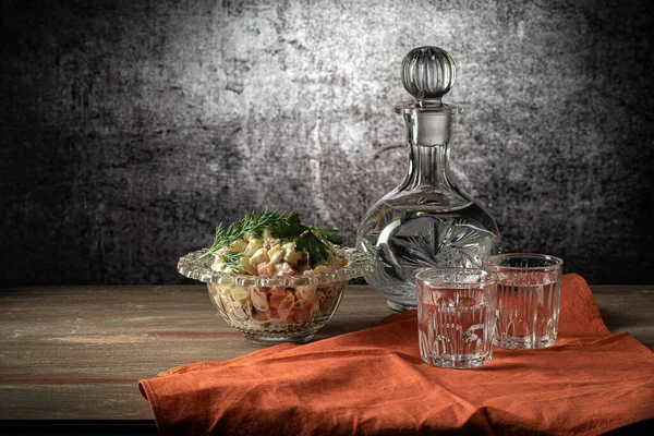 stock image Salad bowl with olivier salad, with glass and decanter, red napkin, on a wooden table top, on a gray background