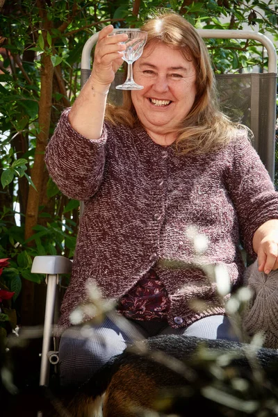 白いワインを飲みながら庭の机の上に座り手で挨拶をする幸せな中年の女性 — ストック写真