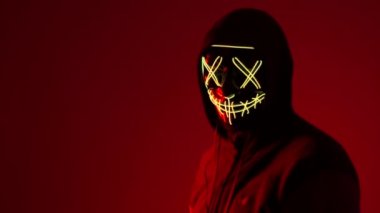 Anonymous kafasını sallıyor, cadılar bayramı maskesi, bilinmeyen korku filmi karakteri, maskeli kötü adam. Kırmızı arka planda, neon ışıklı maskeli bir adam. Kırmızı arka planda neon ışıklı bir adam.
