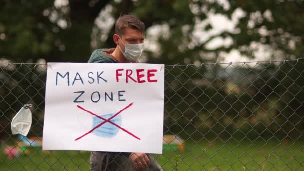 Een poster op het hek met de inscriptie Masker Free Zone. Een jonge mannelijke activist komt naar voren en doet zijn masker af. Ik hing het aan het hek naast de poster. Anti-maskerconcept, covid-denier — Stockvideo