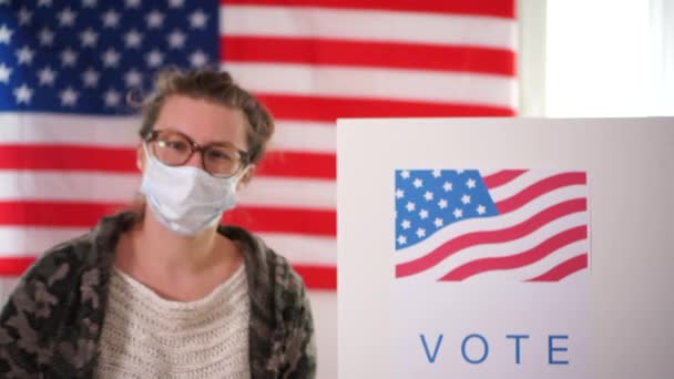 Mujer con máscara médica mostrando que voté por correo con la bandera de los Estados Unidos como fondo. En el fondo, un hombre se acerca a una cabina de votación, nosotros elecciones 2020, covid-19 pandemia coronavirus — Vídeos de Stock