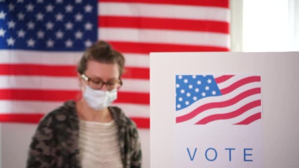 Молодая женщина в маске возле кабины для голосования демонстрирует наклейку. На заднем плане к нам подходит человек, чтобы проголосовать. Выборы в США 2020 года в рамках COVID-19 — стоковое видео