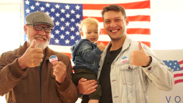 Отец, сын и внук держат наклейки для голосования. Три поколения американской семьи пришли голосовать на избирательном участке. Жест вверх пальцами — стоковое видео