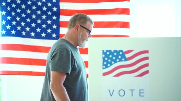 En äldre man med glasögon och grått skägg röstar på en vallokal. Patriot under 2020-talet oss val i röstbåset — Stockvideo