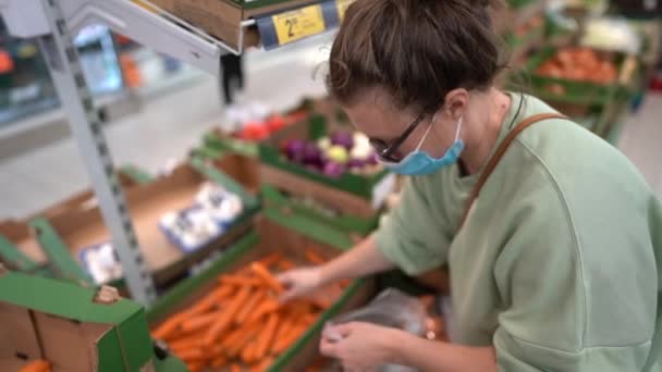 Vrouw met gezichtsmasker en handschoen pikt wortelen op in de groentenafdeling van de supermarkt. Meisje kiezen, op zoek naar kruidenier dingen te kopen op plank tijdens coronavirus crisis of covid-19 uitbraak — Stockvideo