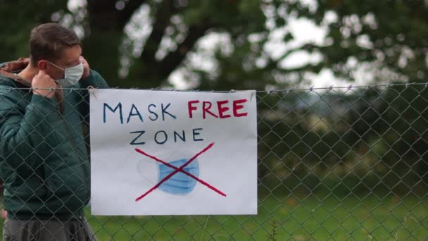 Plakat na ogrodzeniu z napisem Mask Free Zone. Młody aktywista podchodzi i zdejmuje maskę. Powiesiłem go na płocie obok plakatu. Koncepcja anty-maskowa, covid-denier — Wideo stockowe