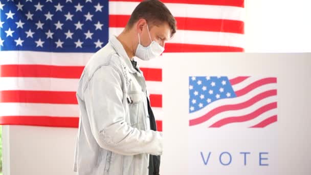 Американец в джинсовой куртке и маске голосует в кабинке для голосования. Выборы в США 2020 года во время пандемии коронавируса ковида-19 — стоковое видео