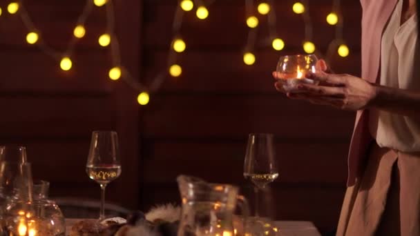Vlak voor de viering: vrouwenhanden zetten een kaars op kerstversierde tafel met kristallen glazen, wijnfles, kaars en kerstverlichting op de achterkant — Stockvideo