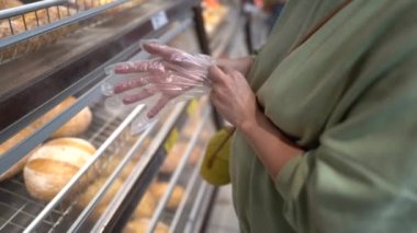 Maske takan genç bir kadın bir süpermarketin ekmek bölümünde tek kullanımlık eldiven takıyor. PPE, kişisel koruyucu ekipman konsepti