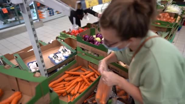 A young woman wearing a mask and protective gloves buys carrots in the vegetables section of a supermarket. Hygiene and precautions concept, covid restrictions during the coronavirus pandemic — Stock Video