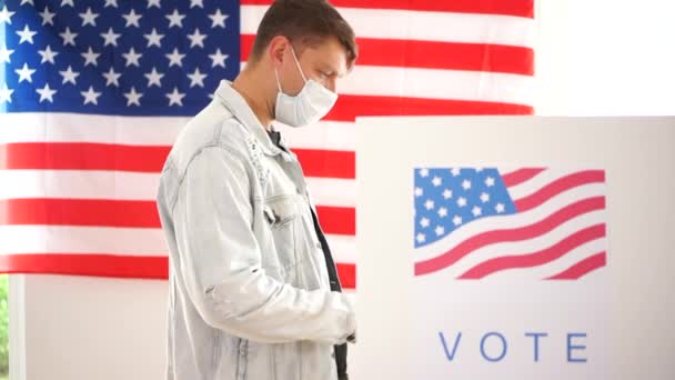 Американський чоловік в джинсовій куртці і маска голосує в кабінці для голосування. Вибори в США 2020 під час коронавірусної пандемії — стокове відео