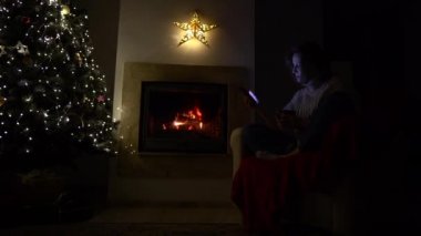 Genç bir kadın şöminenin yanında tek başına oturuyor ve elinde telefon olan bir Noel ağacı var. Sosyal mesafe. Kilitleme sırasında Noel, yeni yıl ve covid-19 koronavirüs salgını